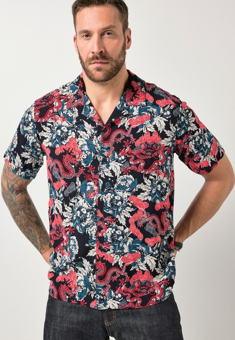 Homme avec barbe et tatouage portant une chemise à manches courtes boutonnée avec des motifs floraux et cachemire rouges, bleus et blancs, debout les mains sur les hanches.