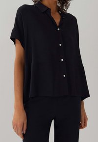 Blusa nera a maniche corte in tessuto leggero, con colletto, chiusura frontale a bottoni e un fit rilassato con orlo a strati.