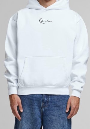 Personne portant un sweat à capuche blanc avec une poche avant et un logo noir en cursive, associé à un jean en denim bleu, sur fond gris.