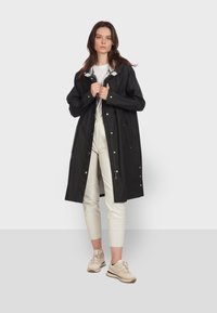 Czarny wodoodporny płaszcz z zatrzaskami, regulowanym kapturem i kieszeniami bocznymi. Połączony z jasnymi spodniami i beżowymi trampkami.