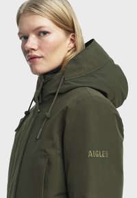 Mladá žena s blond vlasmi, oblečená v olivovo zelenom bundovom kabáte s kapucňou, na ktorom je na rukáve logo AIGLE 1853, otočená mierne doľava.