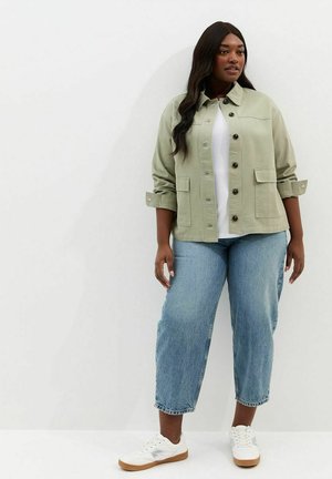 Femme portant une veste verte claire boutonnée, une chemise blanche, un jean bleu raccourci et des baskets blanches, debout contre un mur blanc.