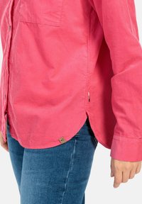 Roze corduroy shirt met lange mouwen, zijsplitten en een borstzak, met een merketiket aan de zoom. Gepaard met blauwe jeans.