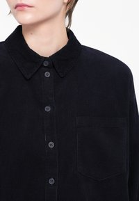 Cămașă din catifea de culoare navy, cu guler, nasturi frontali și un buzunar pe piept. Țesătura are un aspect texturat, cu nervuri.
