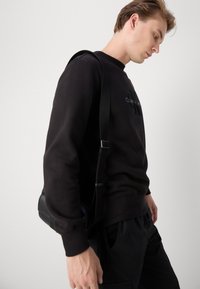 Zwarte sweatshirt van zacht materiaal, met een ronde hals, geribde manchetten en een geborduurd logo op de borst. Model draagt een schoudertas.