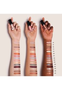 Drei Hände halten glänzende Lippenfarbe-Tuben vor einem neutralen Hintergrund. Armabstriche zeigen verschiedene Farbtöne, die von Nudetönen bis zu tiefen Tönen reichen.