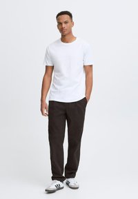 Hvid bomulds t-shirt kombineret med mørke bukser. Modellen bærer sorte og hvide sneakers og står foran en neutral baggrund.