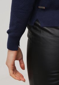 Main détendue sur le côté portant un pull bleu marine texturé et un pantalon en cuir noir avec une petite étiquette métallique de marque sur le pull.