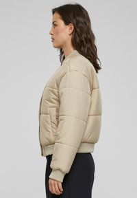 Beige vadderad bomberjacka med ribbad krage och ärmslut, utrustad med en sidficka och en slät, glasyrliknande finish.