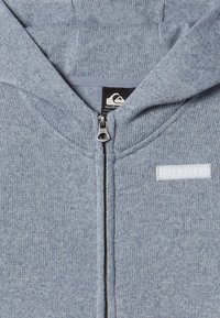 Sudadera gris con cremallera hecha de tela texturizada. Cuenta con una capucha, cremallera metálica y una pequeña etiqueta blanca en el frente.