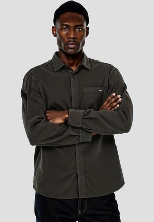 Hombre con camisa oscura de botones y gorro negro, con los brazos cruzados, mirando hacia adelante sobre un fondo liso gris claro.