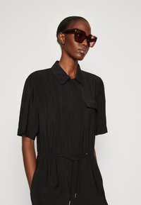 Robe noire à manches courtes avec col, dotée d'une poche avant et d'une taille élastique. Modèle portant des lunettes de soleil rectangulaires sombres. Texture lisse.