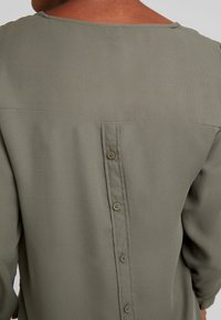 Olivegrünes Blusenshirt mit glatter Textur, rechteckigem Ausschnitt und Knopfverschluss am Rücken.