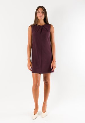 Donna in piedi che indossa un vestito senza maniche viola scuro al ginocchio e tacchi bianchi con punta affilata su sfondo bianco.