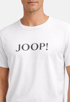 Mies, jolla on valkoinen pyöreäkauluksinen t-paita, jossa musta "JOOP!"-teksti keskellä rintaa.