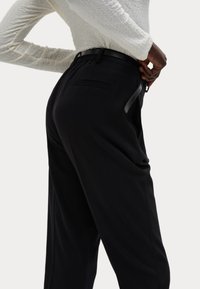 Zwarte broek met een losse fit, zijzakken en een slanke zwarte riem. Zacht materiaal met een lichte glans, weergegeven vanuit een zijaanzicht.