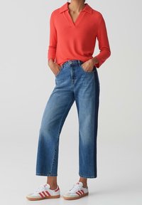 Pull rouge à col, jean bleu ample et baskets blanches à rayures rouges. Détails notables : la texture du pull et la coupe droite du jean.