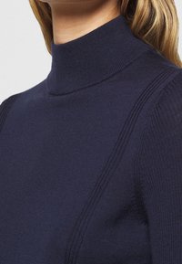 Granatowy sweter z golfem, wykonany z prążkowanej tkaniny, z wysokim kołnierzem i delikatnymi pionowymi wzorami wzdłuż ramion. Gładka faktura.