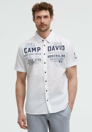 Man with short curly hair wears white short-sleeve button-up shirt with blue printed text, standing with one hand in pocket against plain background.