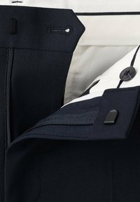 Gros plan de la ceinture d'un pantalon bleu marine montrant la fermeture éclair, le crochet et la patte, le bouton et la doublure en tissu blanc avec un détail de rayure noire.