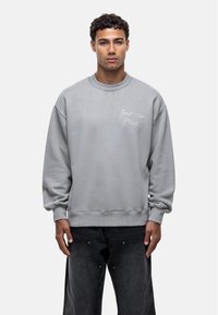 Felpa grigia oversize con colletto e polsini a costine. Presenta testo ricamato sul davanti. Abbinata a jeans scuri.