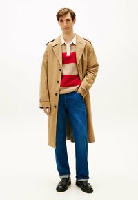 Beżowy trencz nałożony na sweter w czerwono-beżowe paski, w połączeniu z niebieskimi dżinsami i czarnymi butami. Płaszcz ma kołnierz i guziki.