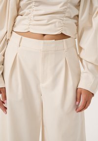 Beige high-waisted trousers with pleats, tailored fit, smooth fabric, and side pockets. Pairing top features gathered texture and long sleeves.