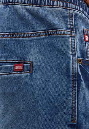 Jasnoniebieskie dżinsy z kontrastującymi pomarańczowymi ściegami. Posiadają czerwoną metkę z logo Big Star na tylnej kieszeni oraz klasyczny design kieszeni.