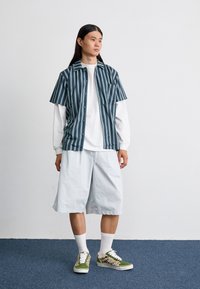 Camisa de manga corta a rayas en tonos azul verdoso y marino, superpuesta a una camiseta de manga larga blanca; combinada con pantalones cortos azul claro y zapatillas con estampado.