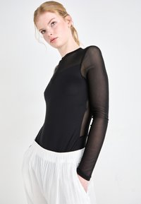 Black bodysuit with long sheer sleeves and a deep V-neck, paired with white, textured, relaxed-fit pants featuring an elastic waistband.