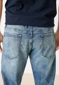 Jeans de mezclilla azul claro con un lavado desgastado, que cuentan con dos bolsillos traseros y costuras visibles. Llevados con una camisa de color marino.