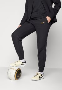 Crne Nike trenirke s dizajnom suženih nogavica, uparene s krem čizmama za nogomet s crnim akcentima, koje se nalaze na teksturiranoj nogometnoj lopti.