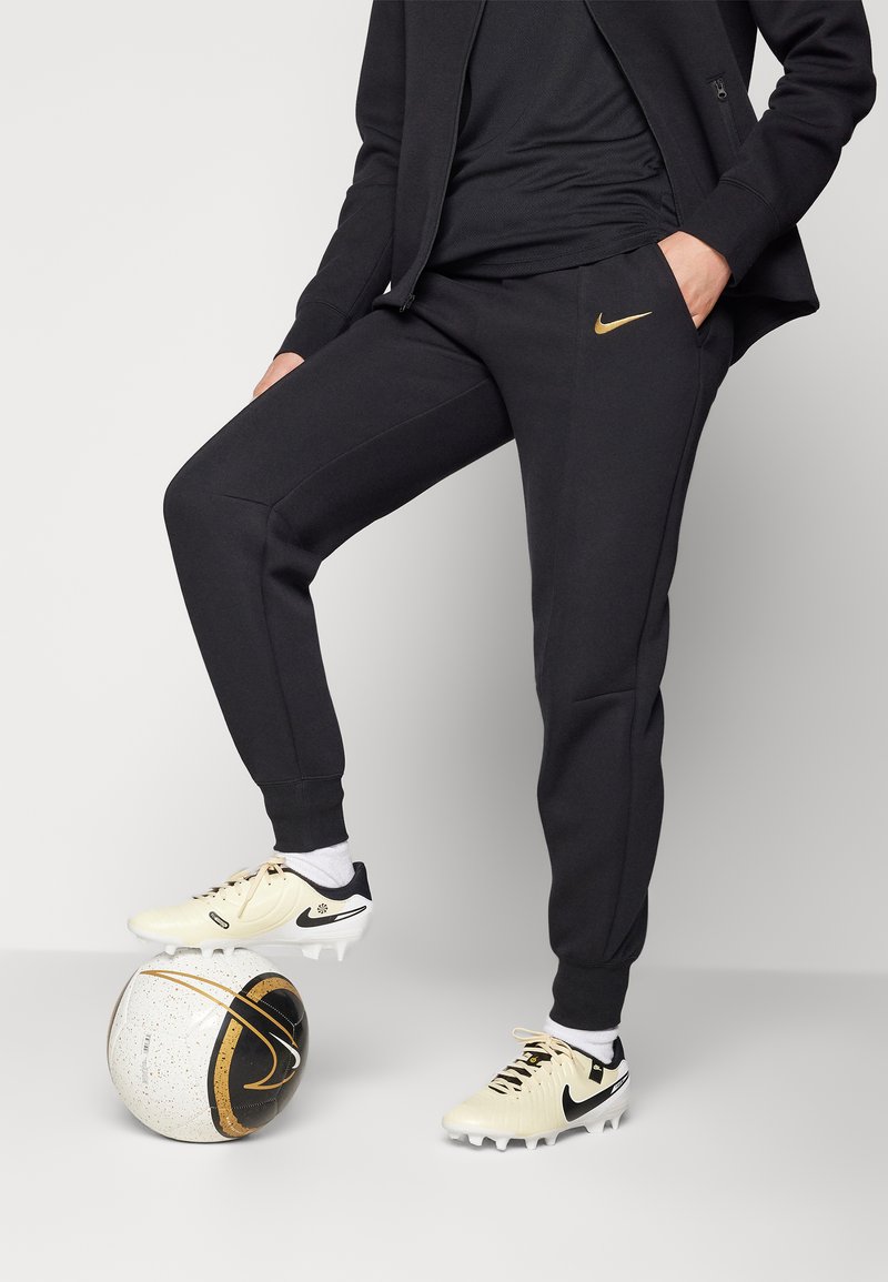 Pantaloni jogger neri Nike con design a gamba affusolata, abbinati a scarpini da calcio crema con dettagli neri, poggiati su una palla da calcio texturizzata.