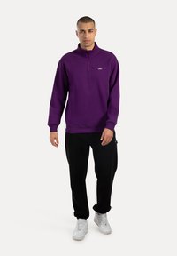 Sudadera de cuello con cremallera de color púrpura, hecha de una mezcla de algodón, con dobladillo y puños de canalé, combinada con pantalones de chándal negros y zapatillas blancas.