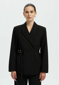 Blazer noir cintré avec un large revers, des manches longues et une ceinture à nouer à la taille. Dispose d'une texture de tissu lisse et d'une silhouette ajustée.