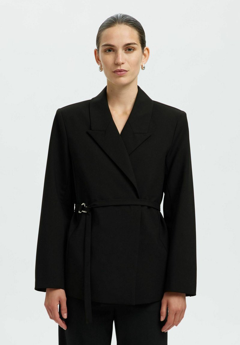 Blazer noir cintré avec un large revers, des manches longues et une ceinture à nouer à la taille. Dispose d'une texture de tissu lisse et d'une silhouette ajustée.