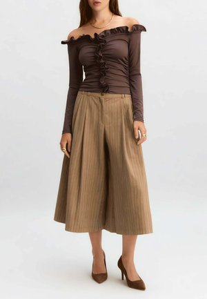 Donna che indossa un top marrone con spalle scoperte e volant, pantaloni beige a gamba larga cropped a righe sottili, tacchi marroni e accessori dorati.