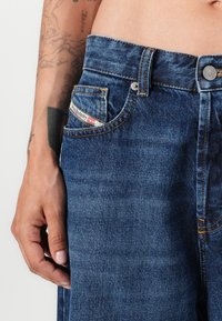 Jeans de denim azul com corte reto, apresentando costuras em contraste, uma pequena etiqueta de bolso e um fecho de botão metálico na cintura.