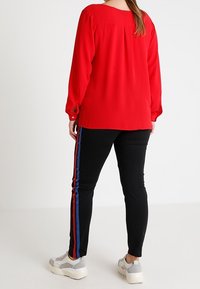 Kobieta ubrana w jasnoczerwoną bluzkę z długim rękawem, czarne spodnie z czerwonymi i niebieskimi paskami po bokach oraz białe trampki, odwrócona plecami do kamery.