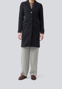 Personne portant un manteau noir boutonné, un pantalon gris à jambes larges et des mocassins marron, debout devant un fond uni gris clair.