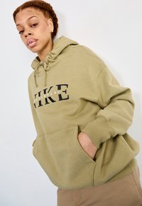 Alyvuogių spalvos džemperis su vilkimo virve, priekinė kišenė ir juodu tekstu "NIKE" ir "SPORTSWEAR" atspausdintu krūtinėje. Minkštas medvilnės audinys.
