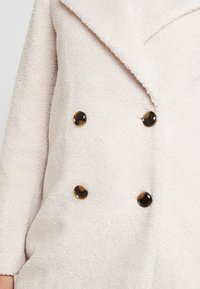 Jasno beżowy dwurzędowy płaszcz wykonany z teksturowanej plecionki, z dużymi ciemnymi guzikami w kolorze żółwia i szerokim kołnierzem.