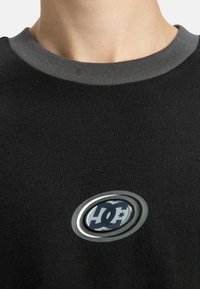 T-shirt en coton noir avec un col en maille gris, arborant un logo circulaire en bleu et argent au centre, créant un contraste audacieux.