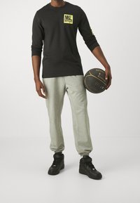 Maglietta nera a maniche lunghe con logo Nike giallo, pantaloni sportivi grigi chiari con orli, e scarpe da basket nere, che tiene in mano un pallone da basket nero.
