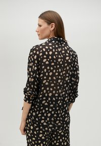 Sheer black blouse with cream polka dots, long sleeves, and a rolled cuff, featuring a back button closure and a relaxed fit.