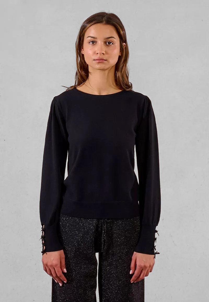 Donna con capelli castani lisci, che indossa un top nero a maniche lunghe e pantaloni scuri brillanti, in piedi contro uno sfondo grigio.