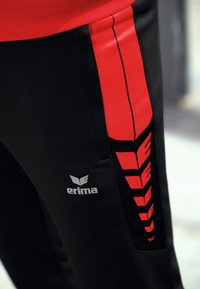 Schwarze Sporthose mit einem roten Streifen an der Seite, versehen mit einem weißen Logo "erima" an dem Unterschenkel.