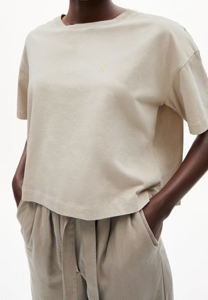 Vaaleanbeige lyhythihainen t-paita, joka on valmistettu pehmeästä puuvillasta ja jossa on pieni kultainen logo rinnassa. Yhdistettynä väljään, vaaleanbeigeen housupariin.