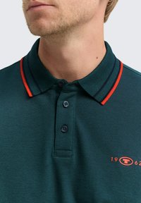 Nahaufnahme eines Mannes, der ein dunkelgrünes Poloshirt mit einem rot-schwarz gestreiften Kragen und rotem "1962"-Logo auf der Brust trägt.