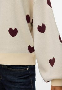 Pull beige avec des motifs de cœurs bordeaux, doté d'un ourlet côtelé et de manches amples, associé à un jean en denim foncé.